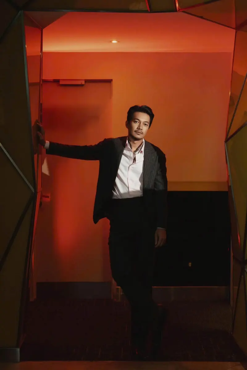 Quang Tuấn in a pinstripe suit and pale shirt, leaning in a red-lit doorway