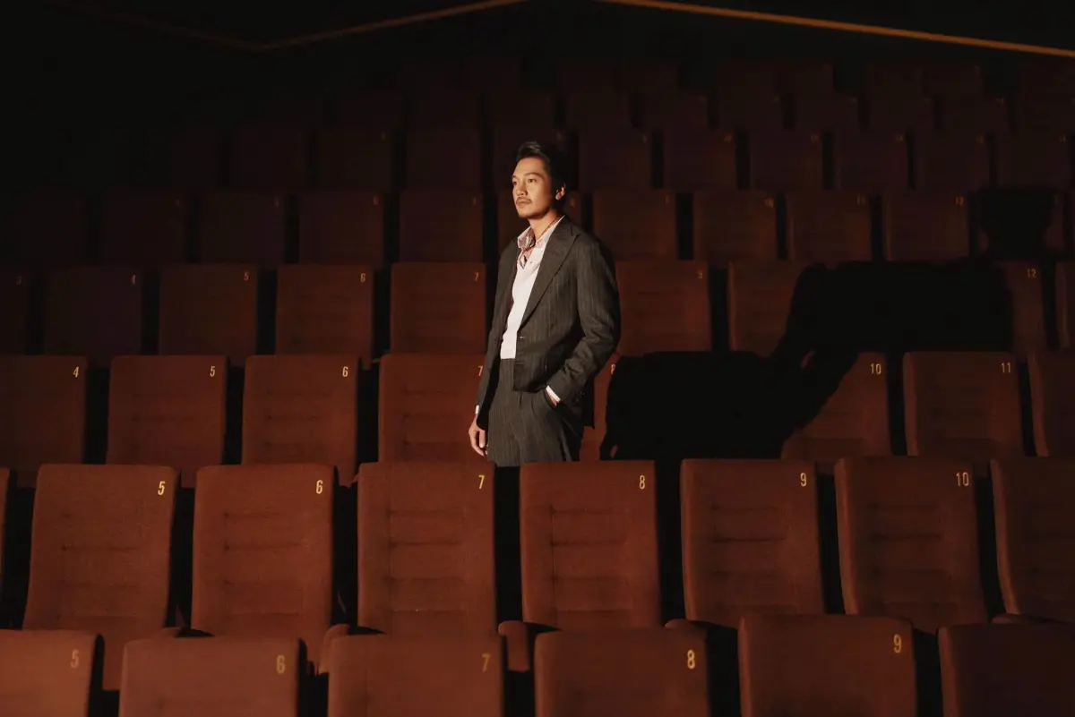 Quang Tuấn in a dark pinstripe suit, standing among rows of cinema seats