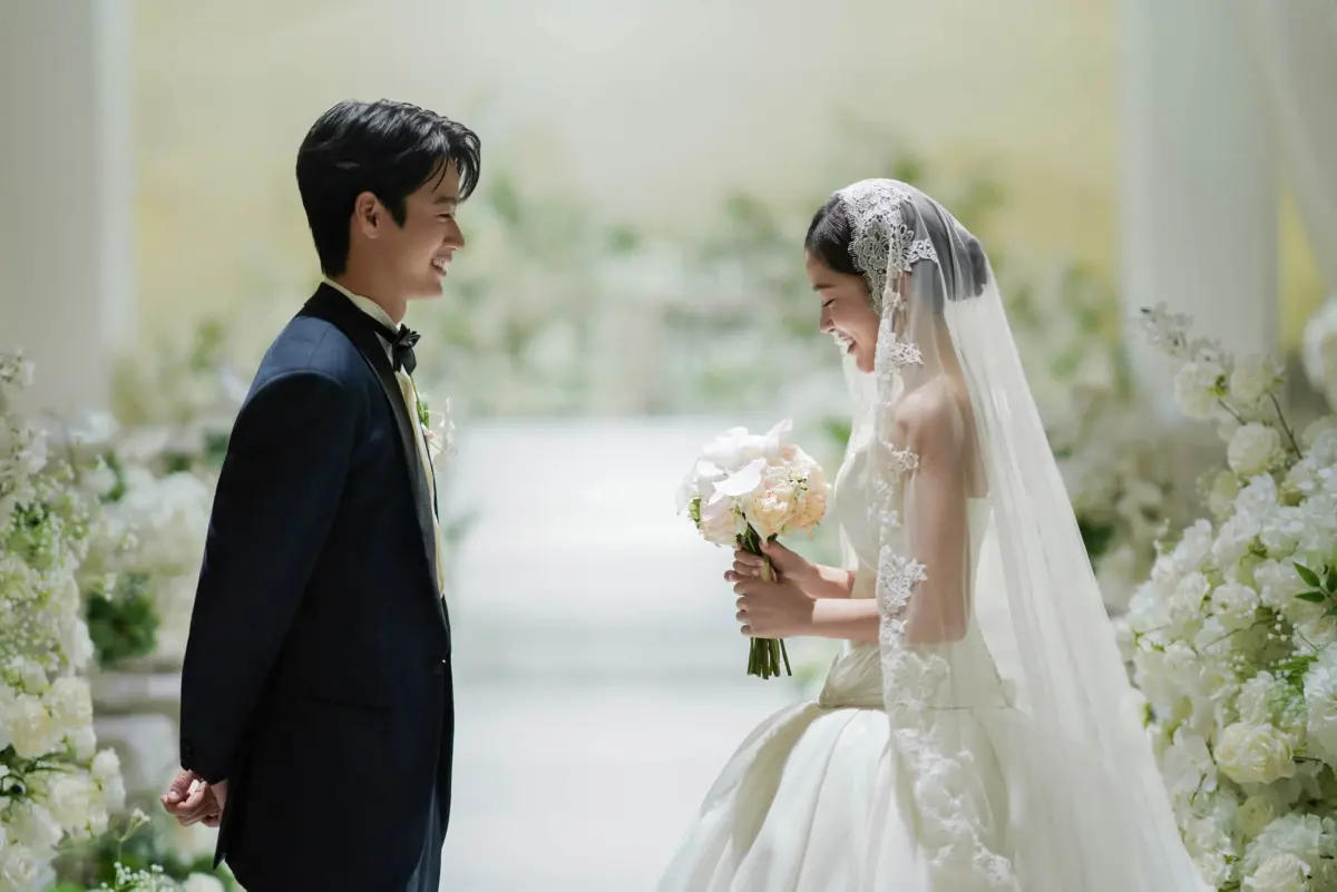 A groom in a dark tuxedo and a bride in a veil facing each other among white flowers