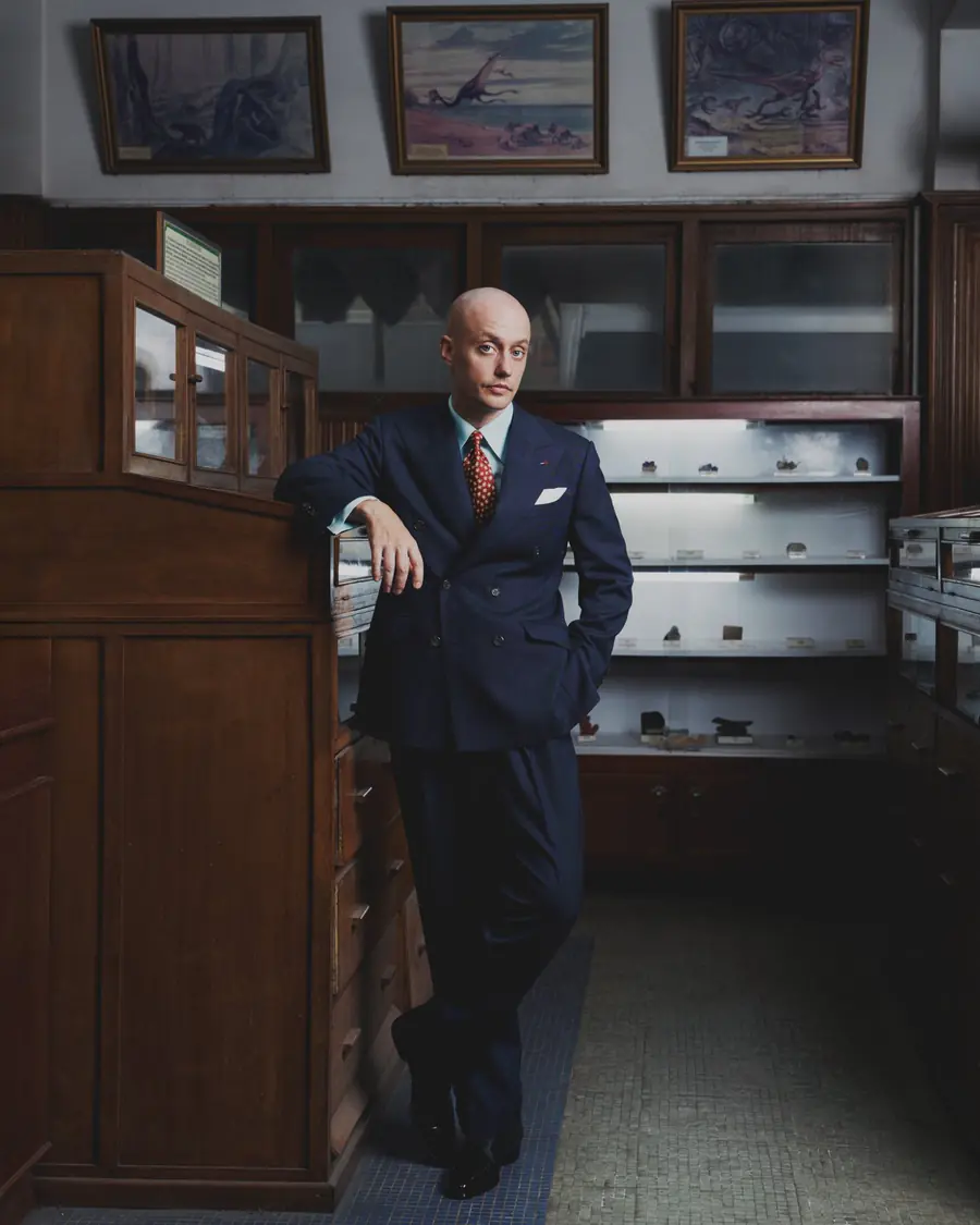 A man in a navy double-breasted suit standing in a wood-panelled tailoring room