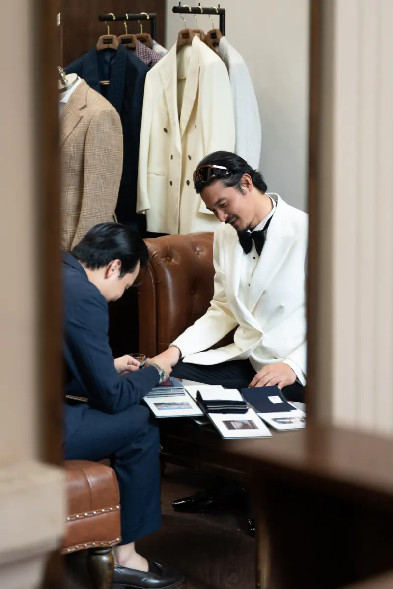 Consultation in the atelier: a client in a white dinner jacket looks through cloth samples with an advisor